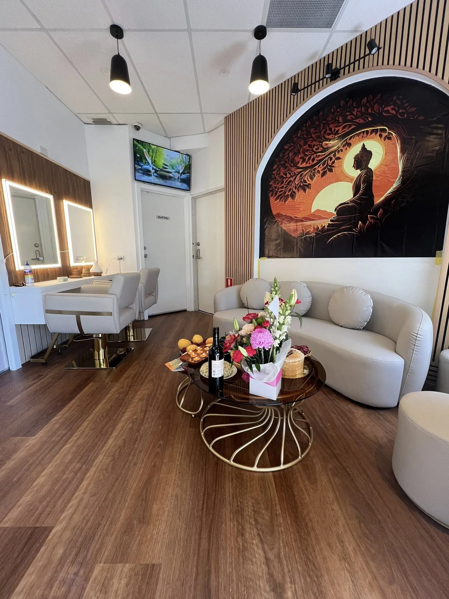 The salon floor — two gold-based styling chairs at lit vanity mirrors on the left, the arched Buddha mural and curved cream sofa on the right, walnut floor