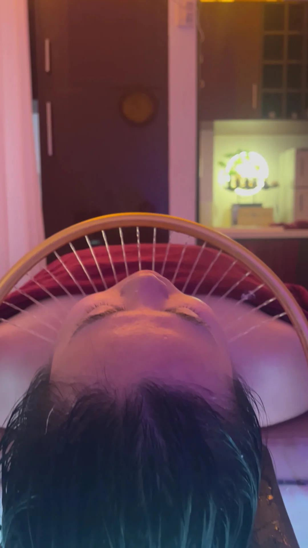 Overhead of a client's face and wet hair beneath the gold arc rinse ring, lit in warm pink-violet light