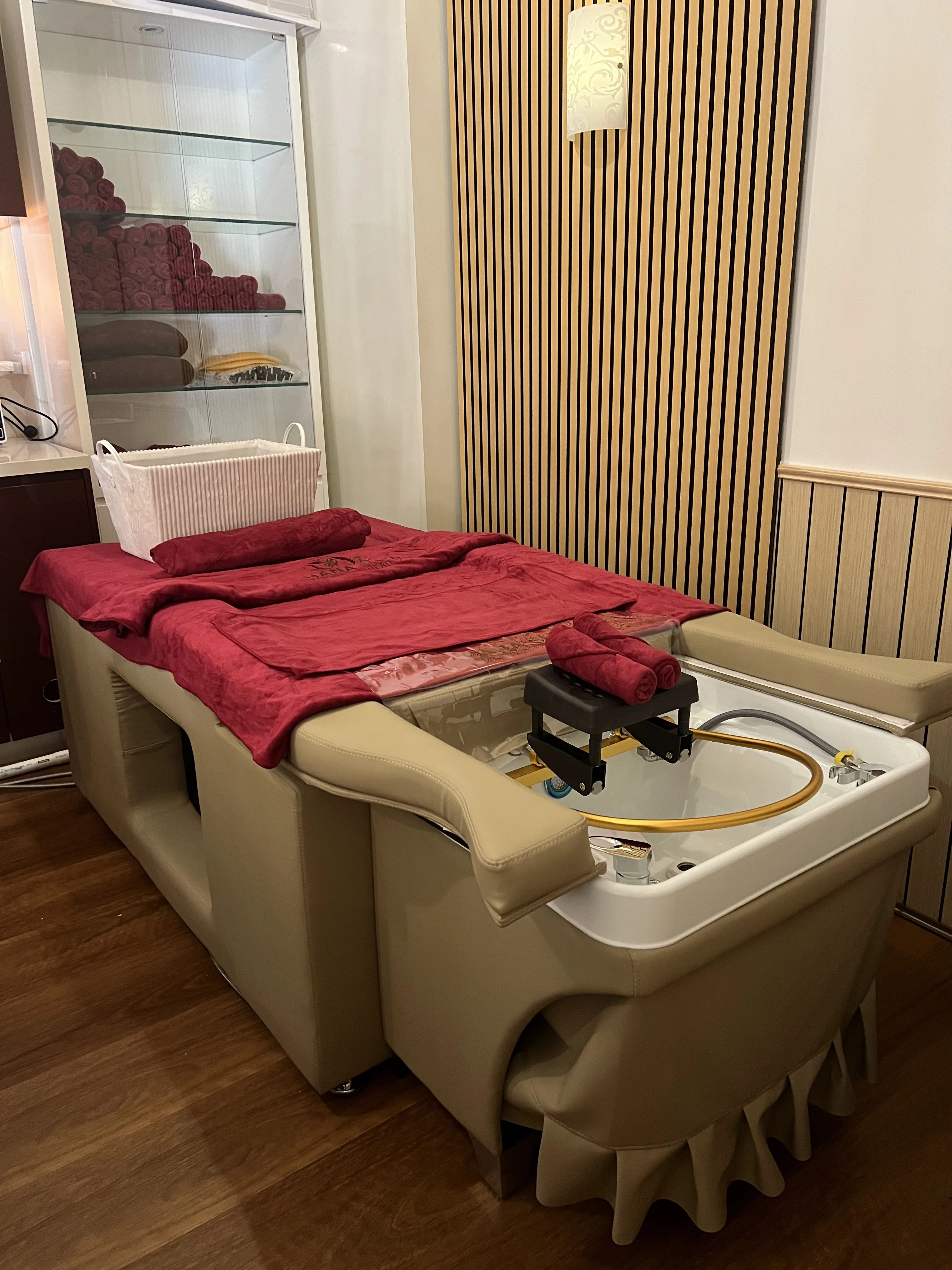 A single head-spa station dressed for a session — cream leather recliner, white basin with gold arc ring, deep-garnet drape and rolled towels, glass towel cabinet behind