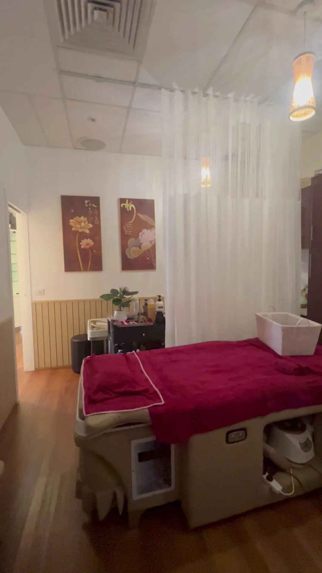 A curtained treatment bay dressed in burgundy, lotus panels on the wall, sheer white curtain, corridor running to the left