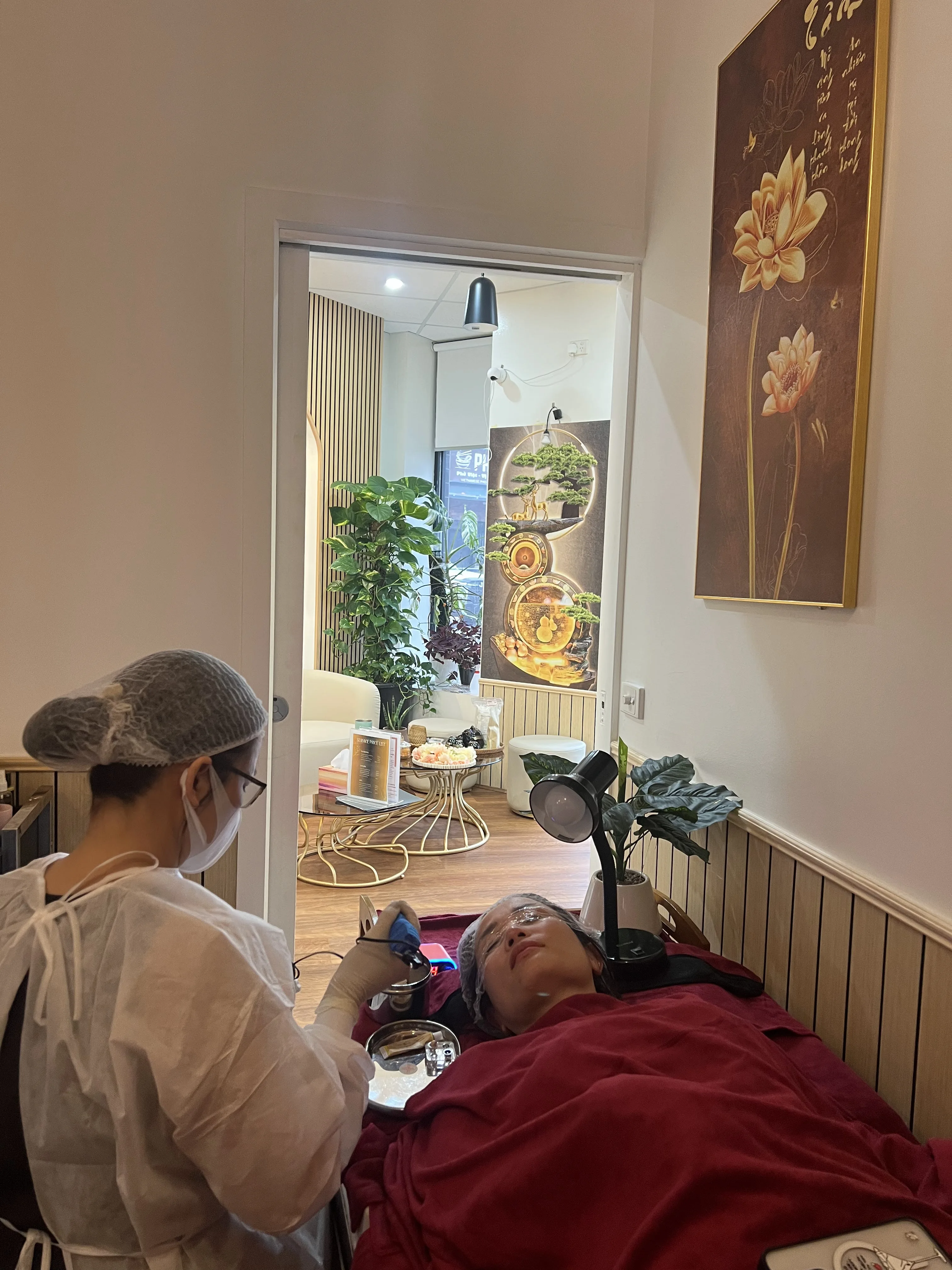 A therapist in white gown and mask performing a facial on a reclined client, lit lounge and gold-toned wall panel visible through the doorway