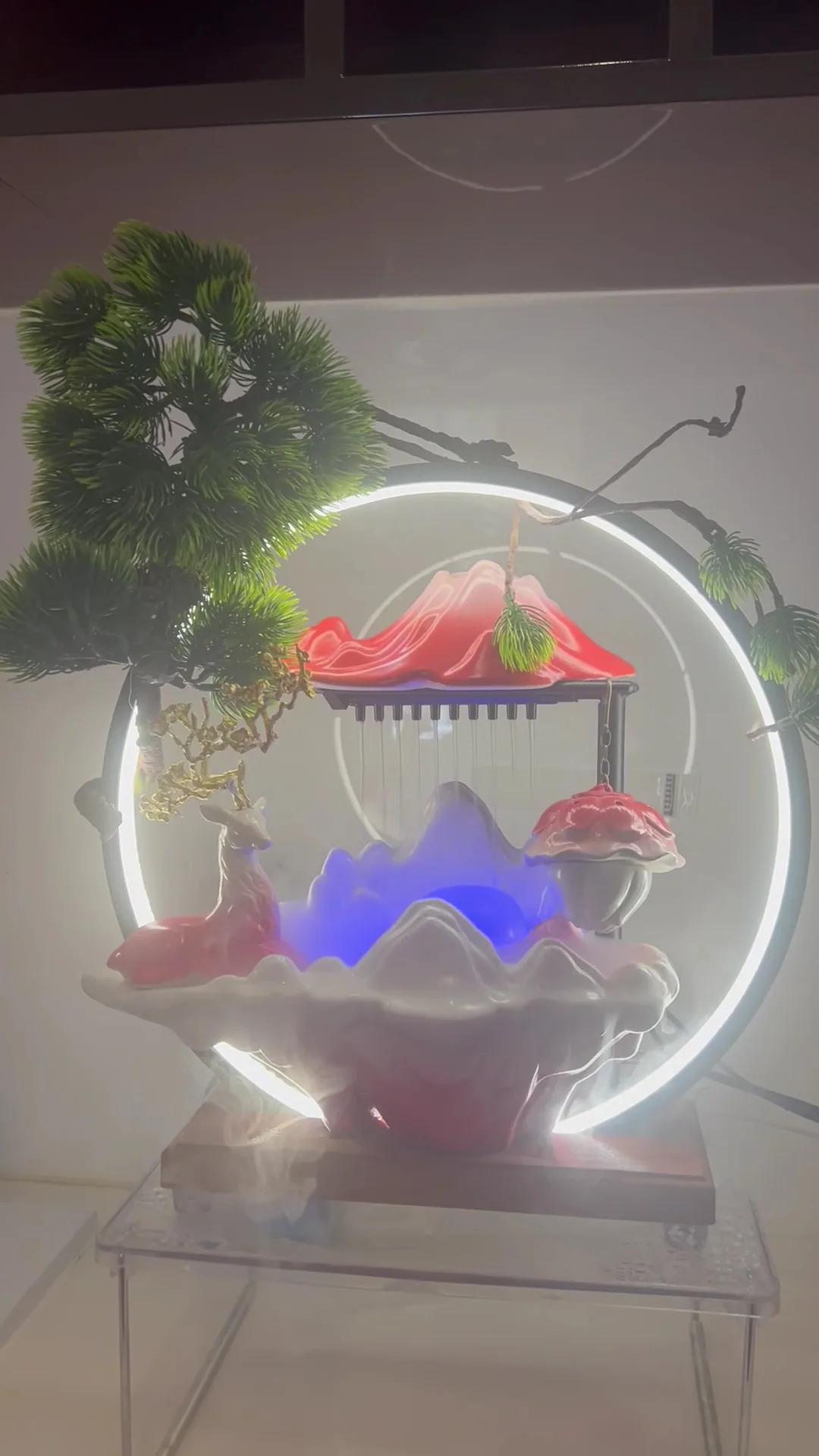 A lit ornamental water feature — ceramic mountains, a deer figurine, bonsai sprigs and a mist plume on an acrylic side table