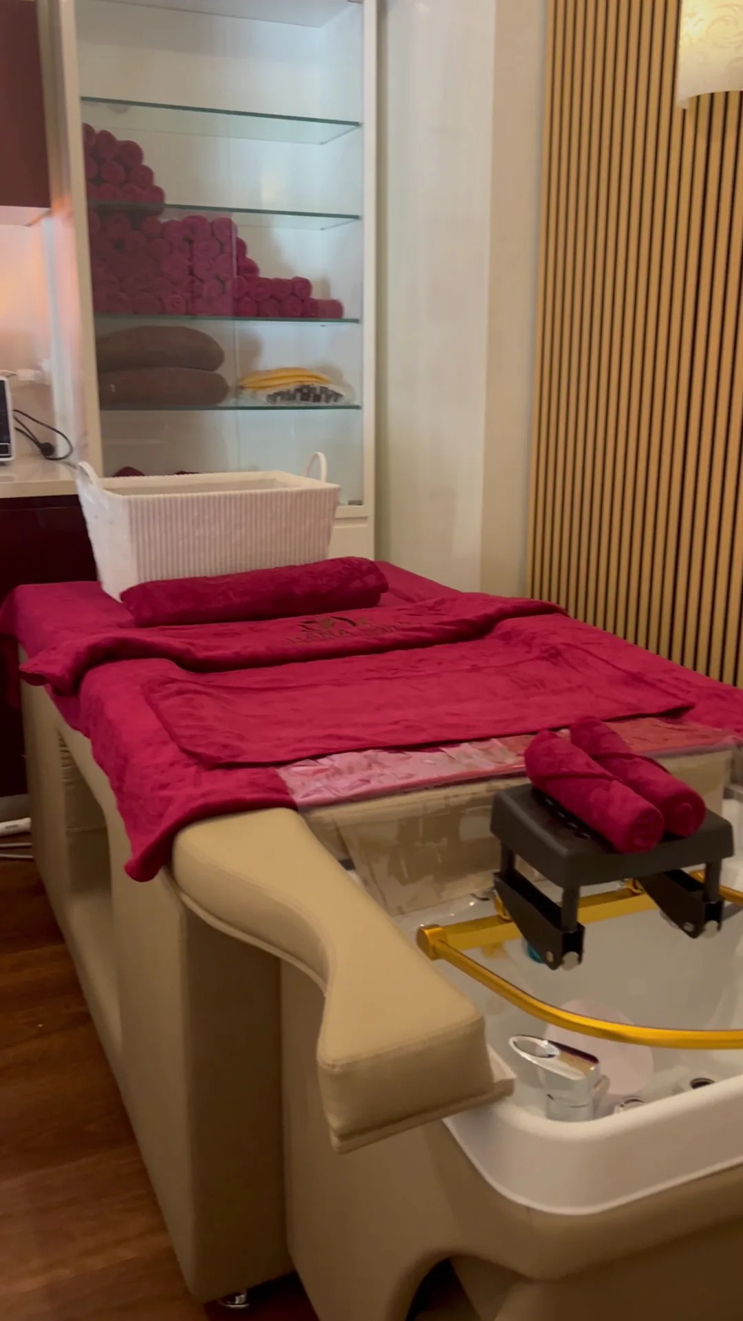A made-up head-spa station in warm lamplight — burgundy embroidered drape and rolled towels on the recliner, gold arc ring over the basin, glass cabinet of rolled burgundy towels behind