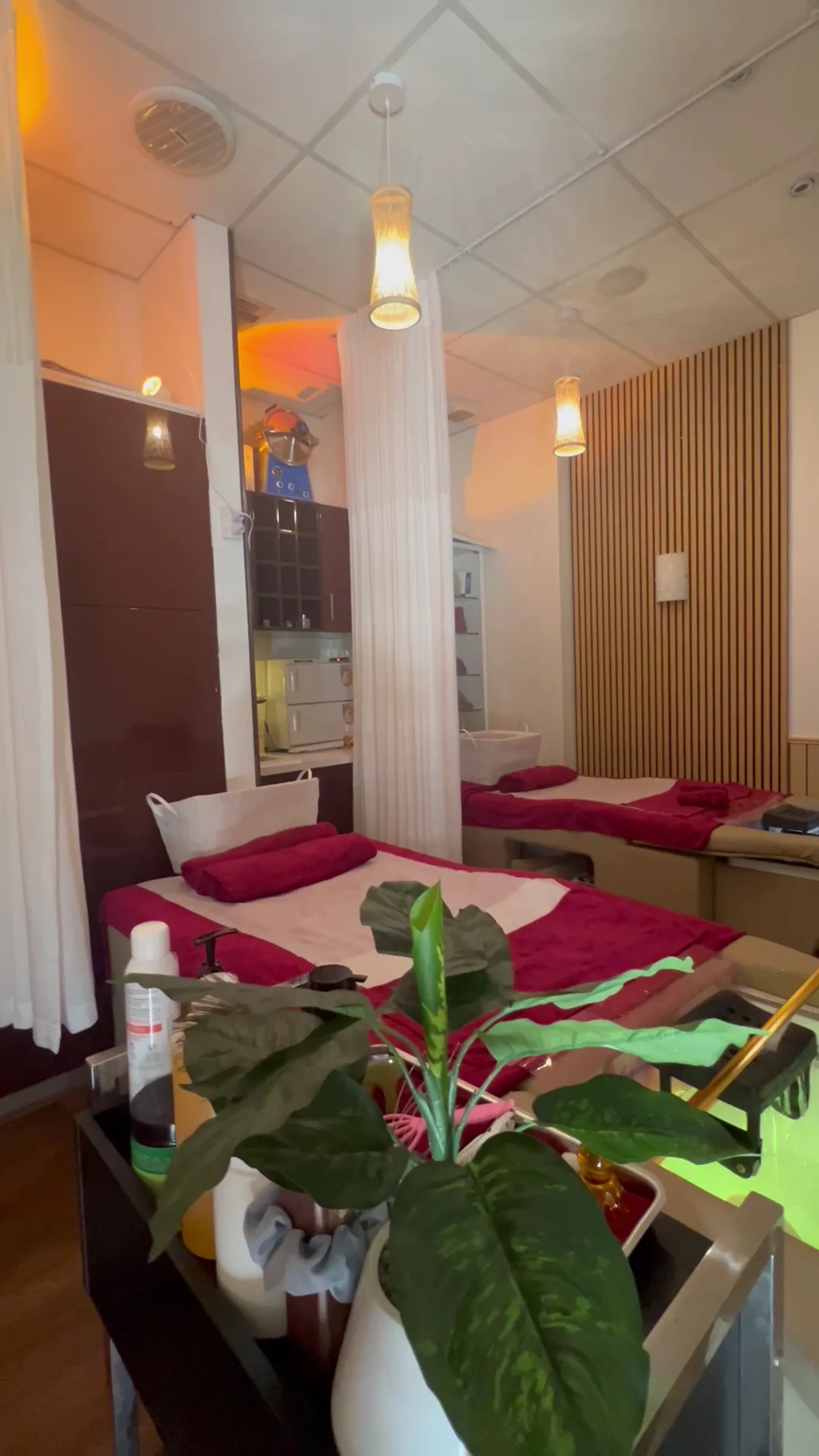 Two curtained treatment bays in warm light — both beds dressed burgundy over cream, woven pendant lamps above, oak batten wall at right