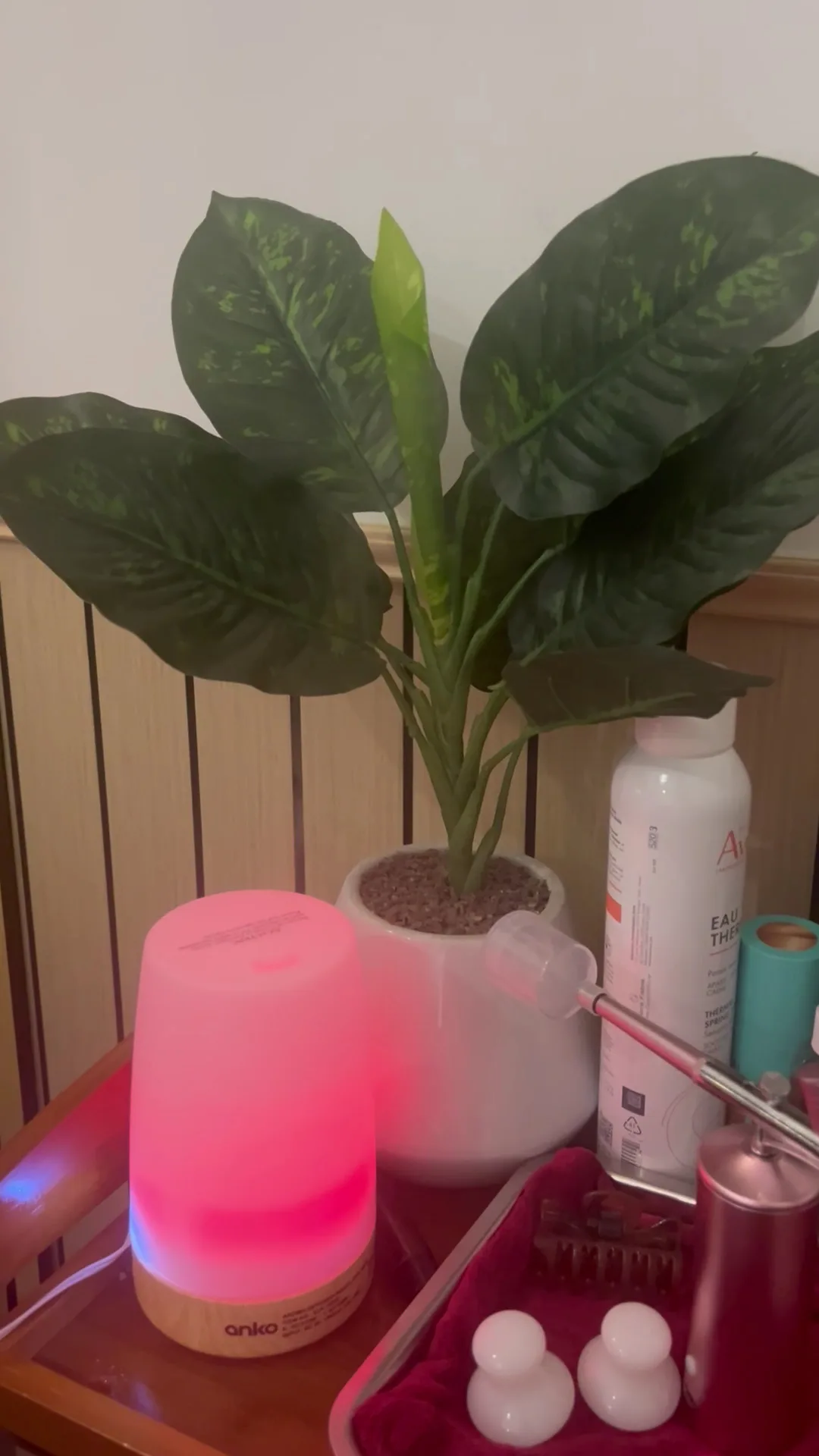 An anko aroma diffuser glowing pink beside a potted plant, Avène thermal spray and an airbrush handpiece on a burgundy-lined tray