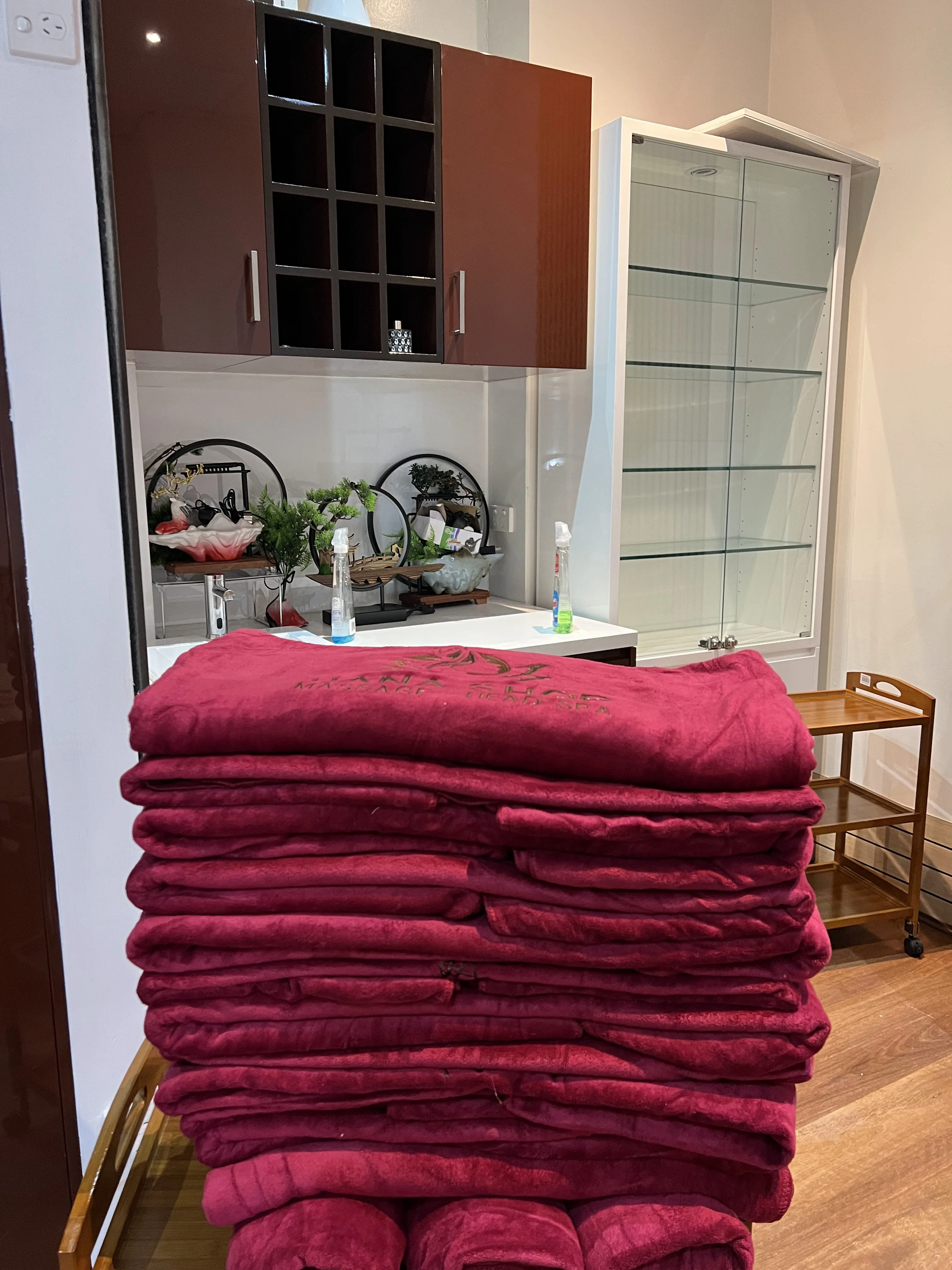 Tall folded stack of deep-garnet microfibre towels on a trolley at the treatment station — gold-thread embroidery on the top towel, dark-red overhead cabinets behind