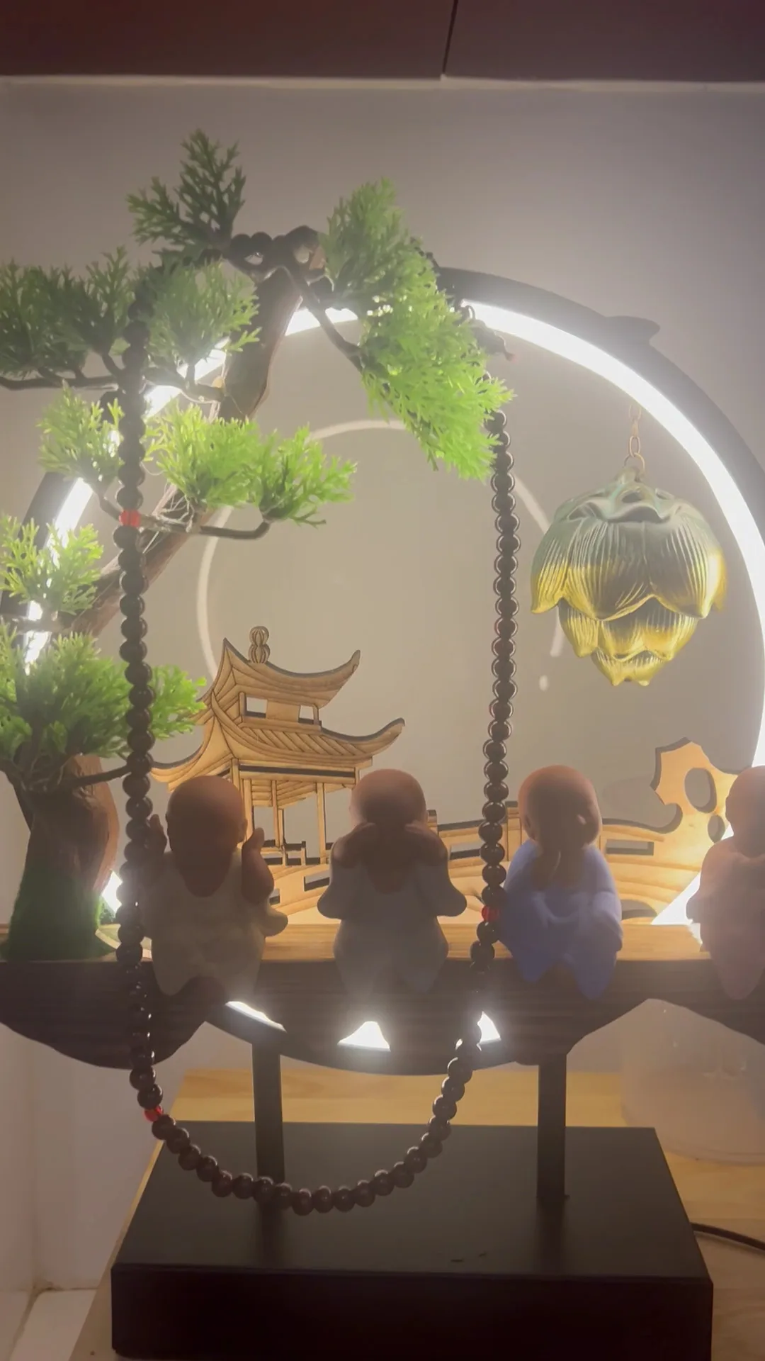 A backlit ring ornament with four small monk figurines, a carved pagoda, bonsai and a hanging lotus lamp